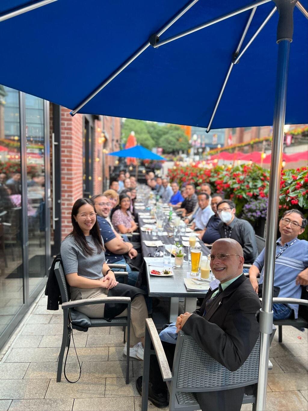 A group enjoys lunch with patio seating around a long outdoor dining table under blue umbrellas, with food, drinks, and flowers adorning the private event.