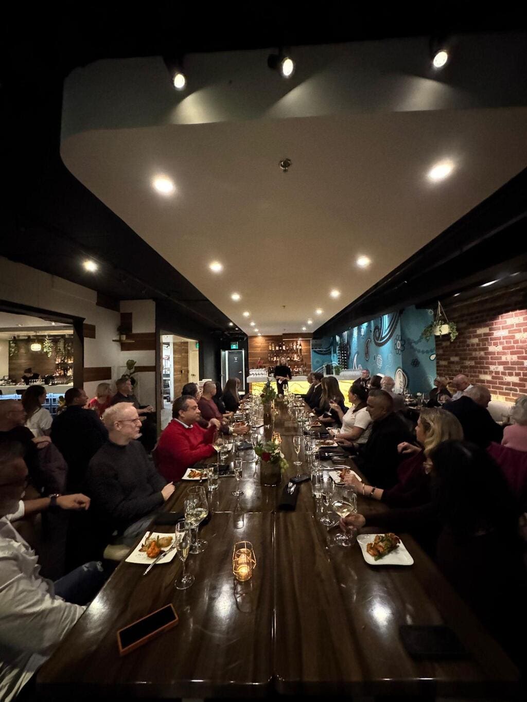 A large group of people sits along a long wooden dining table in a dimly lit restaurant, engaged in conversation and eating dinner—an ideal scene for networking events or as a cocktail party venue that Toronto prides itself on.