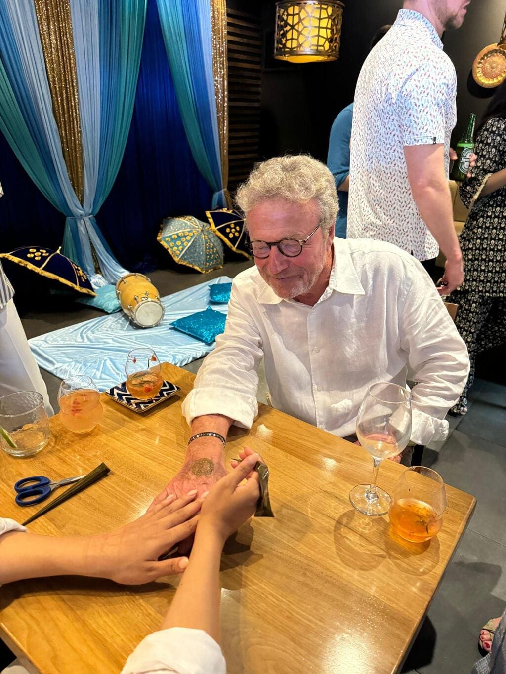 An older man sits at a table in a private room in an Indian restaurant, having his palm read with drinks on the table and decorative blue drapes and props in the background.