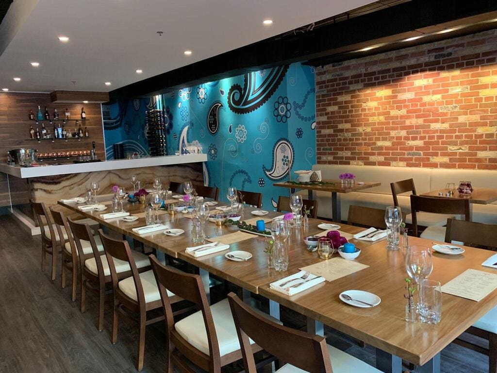 A modern restaurant dining area with a long set table, wooden chairs, brick and blue patterned walls, and a bar in the background—perfect for private events or Special occasion event planning.